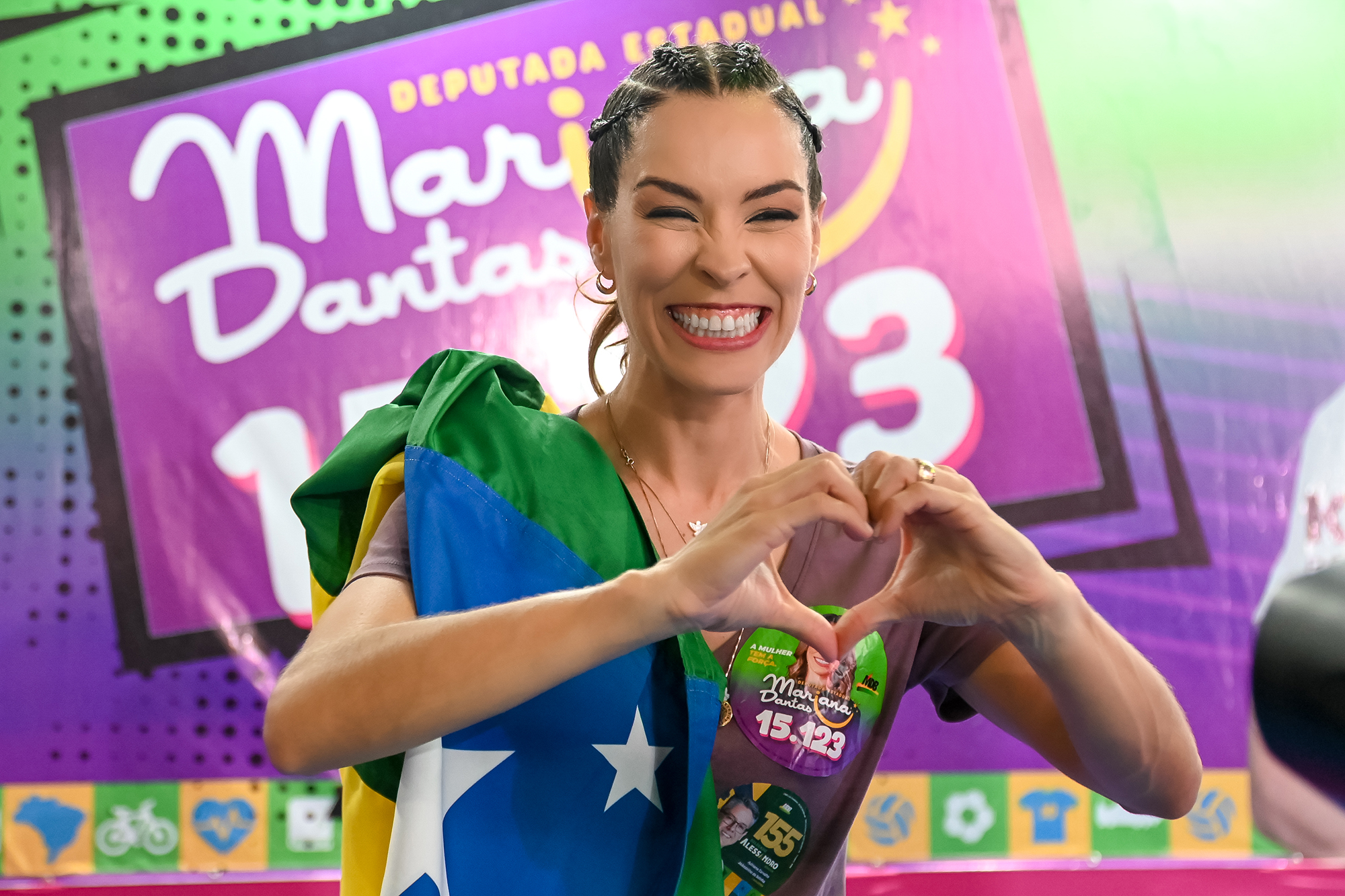 Mariana Dantas fazendo coração com as mãos diante do painel da campanha